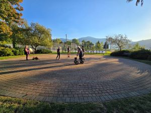 Photograph of an outdoor circular brick labyrinth surrounded by grass, trees, and mountains under a partly cloudy sky. Several people are walking the labyrinth