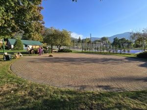 Photograph of an outdoor circular brick labyrinth surrounded by grass, trees, and mountains under a partly cloudy sky. Several people are gathered at the entrance of the labyrinth