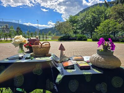 Two alters draped in a dark blue and green cloth with two vases of flowers, assorted rituals objects and a wicker basket. In the background is a circular brick labyrinth with trees and mountains and blue sky with clouds 