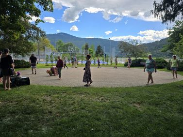Photograph of an outdoor circular brick labyrinth surrounded by grass, trees, and mountains under a partly cloudy sky. Several people are walking the labyrinth