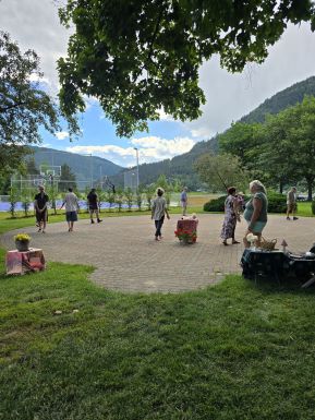 Photograph of an outdoor circular brick labyrinth surrounded by grass, trees, and mountains under a partly cloudy sky. Several people are walking the labyrinth 