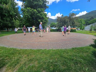 Photograph of an outdoor circular brick labyrinth surrounded by grass, trees, and mountains under a partly cloudy sky. Several people are walking the labyrinth