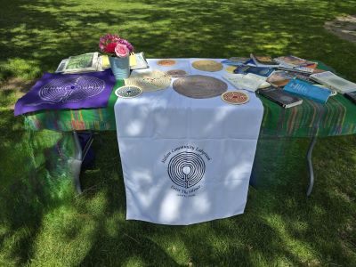 Photograph of an outdoor informational table set up on grass, featuring a white cloth with a circular labyrinth logo and text reading 