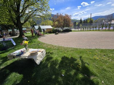 Photograph of an outdoor park area featuring a circular paved labyrinth surrounded by grass and trees under a partly cloudy sky. A small table with flowers, plates, and utensils is set on the grass in the foreground, while benches and a fence are visible in the background near a mountainous landscape. 