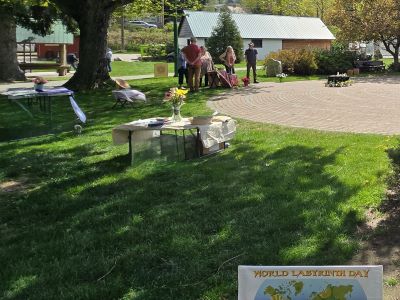 Photograph of an outdoor park setting featuring a circular labyrinth pathway with a group of people standing nearby. A table with flowers and informational materials is set up on the grass, along with a sign reading 