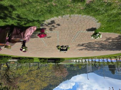 Photograph of an outdoor memorial setup on a circular brick-paved labyrinth surrounded by grass and trees, with mountains and a basketball court visible in the background. The memorial features several flower arrangements placed on the ground and on a small table covered with a pink cloth, indicating a tribute or remembrance event.
