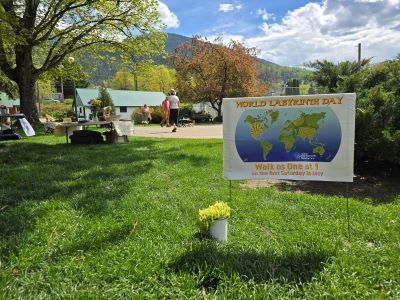 Photograph of an outdoor park setting featuring a sign promoting World Labyrinth Day with a colorful world map and text encouraging participation in a walk event. The scene includes green grass, trees, a few people walking, and a mountainous background under a partly cloudy sky. 