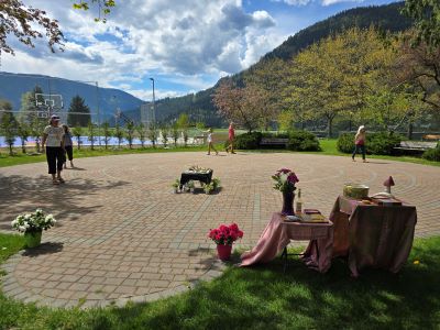 Photograph of an outdoor circular paved labyrinth surrounded by grass, trees, and mountains under a partly cloudy sky. Several people are walking around, with tables covered in pink cloth displaying flowers and decorative items in the foreground, suggesting a casual gathering or event. 