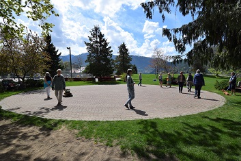 Photograph of a group of people walking and standing on a large circular paved labyrinth in a park surrounded by grass and trees. Background features mountains under a partly cloudy sky, suggesting a peaceful outdoor setting for leisure or exercise.